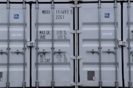 A close-up view of the doors of a shipping container. The surface is metallic with vertical reinforcement bars and stickers indicating information about weight in kilograms and pounds. Labels such as 'MAX. GR. TARE' and 'NET CU. CAP.' are present along with numerical values. The container has a standard industrial appearance.