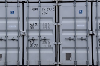 A close-up view of the doors of a shipping container. The surface is metallic with vertical reinforcement bars and stickers indicating information about weight in kilograms and pounds. Labels such as 'MAX. GR. TARE' and 'NET CU. CAP.' are present along with numerical values. The container has a standard industrial appearance.