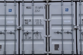 A close-up view of the doors of a shipping container. The surface is metallic with vertical reinforcement bars and stickers indicating information about weight in kilograms and pounds. Labels such as 'MAX. GR. TARE' and 'NET CU. CAP.' are present along with numerical values. The container has a standard industrial appearance.