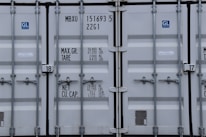 A close-up view of the doors of a shipping container. The surface is metallic with vertical reinforcement bars and stickers indicating information about weight in kilograms and pounds. Labels such as 'MAX. GR. TARE' and 'NET CU. CAP.' are present along with numerical values. The container has a standard industrial appearance.