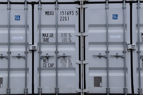 A close-up view of the doors of a shipping container. The surface is metallic with vertical reinforcement bars and stickers indicating information about weight in kilograms and pounds. Labels such as 'MAX. GR. TARE' and 'NET CU. CAP.' are present along with numerical values. The container has a standard industrial appearance.