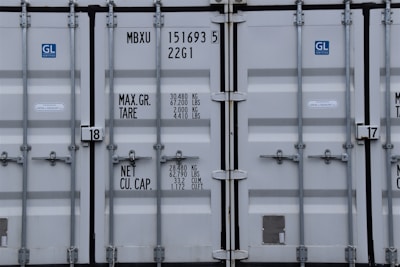 A close-up view of the doors of a shipping container. The surface is metallic with vertical reinforcement bars and stickers indicating information about weight in kilograms and pounds. Labels such as 'MAX. GR. TARE' and 'NET CU. CAP.' are present along with numerical values. The container has a standard industrial appearance.