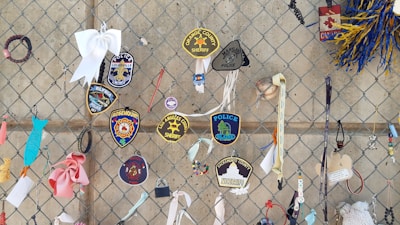 A variety of police and sheriff patches are attached to a chain-link fence. The patches include names like Louisville, Orange County, and Los Angeles County. There are also ribbons, wristbands, and small ornaments, including a white bow and a small baseball.