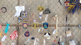 A variety of police and sheriff patches are attached to a chain-link fence. The patches include names like Louisville, Orange County, and Los Angeles County. There are also ribbons, wristbands, and small ornaments, including a white bow and a small baseball.