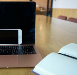 A laptop with a black screen is placed on a light wooden table with an open notebook on one side and a smartphone resting against the laptop. The room has a modern interior with chairs in the background.