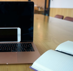 A laptop with a black screen is placed on a light wooden table with an open notebook on one side and a smartphone resting against the laptop. The room has a modern interior with chairs in the background.