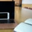 A laptop with a black screen is placed on a light wooden table with an open notebook on one side and a smartphone resting against the laptop. The room has a modern interior with chairs in the background.