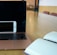 A laptop with a black screen is placed on a light wooden table with an open notebook on one side and a smartphone resting against the laptop. The room has a modern interior with chairs in the background.