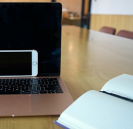 A laptop with a black screen is placed on a light wooden table with an open notebook on one side and a smartphone resting against the laptop. The room has a modern interior with chairs in the background.