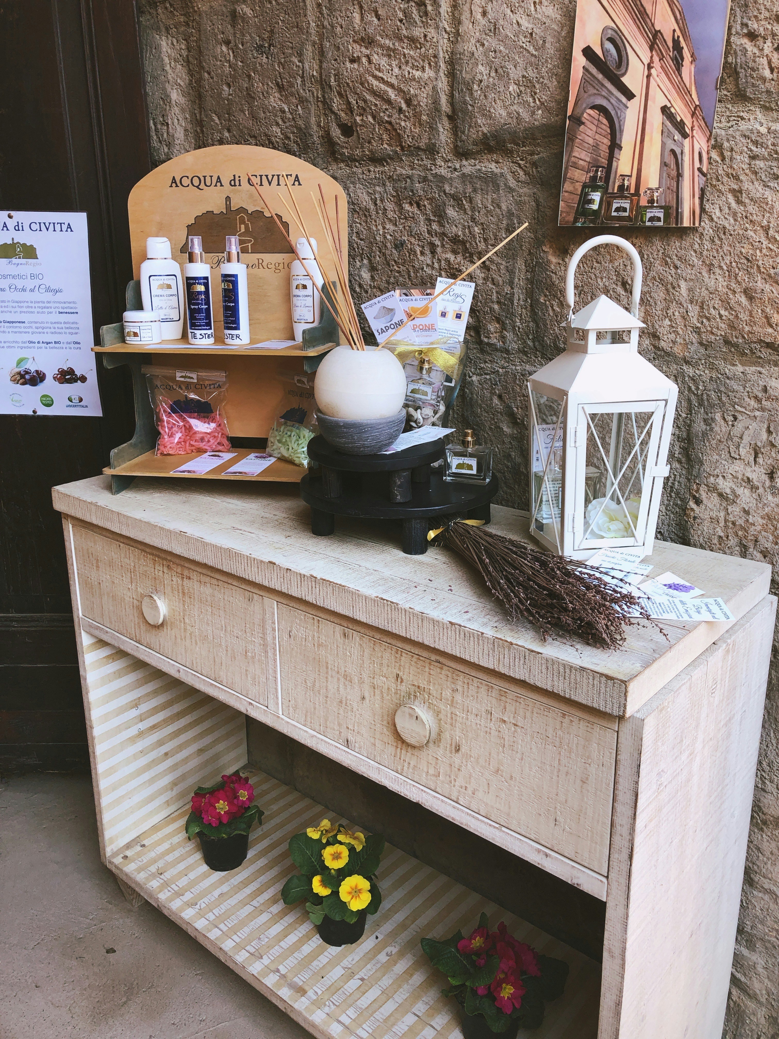 A rustic wooden shelf holds a collection of spa and wellness products, including bottles and packages labeled 'Acqua di Civita'. A white lantern and a bunch of dried lavender are placed on the right side. Below, colorful potted flowers are displayed on the shelves.