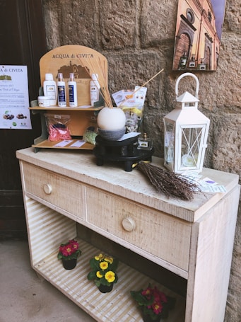 A rustic wooden shelf holds a collection of spa and wellness products, including bottles and packages labeled 'Acqua di Civita'. A white lantern and a bunch of dried lavender are placed on the right side. Below, colorful potted flowers are displayed on the shelves.
