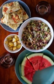 A variety of dishes are arranged on a wooden table. There is a plate of flatbread topped with tomatoes and herbs, a bowl of mixed bean salad with leafy greens, a small bowl of roasted potatoes, and a plate of watermelon slices. Two glasses, presumably filled with a beverage, are also present.