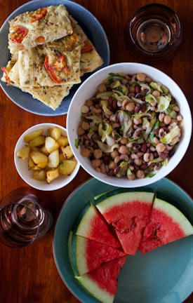 A variety of dishes are arranged on a wooden table. There is a plate of flatbread topped with tomatoes and herbs, a bowl of mixed bean salad with leafy greens, a small bowl of roasted potatoes, and a plate of watermelon slices. Two glasses, presumably filled with a beverage, are also present.