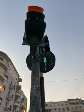 A traffic light positioned against the backdrop of a clear sky. The light is green with an amber colored visor at the top. Surrounding buildings have multiple floors and balconies, adding an urban feel to the scene.