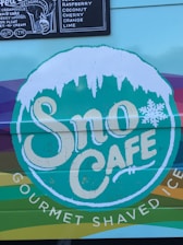 A colorful sign for Sno Cafe is displayed with a snow-like design on top of the lettering. There is also a menu board listing various ice cream flavors such as raspberry, coconut, cherry, and lime.
