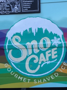 A colorful sign for Sno Cafe is displayed with a snow-like design on top of the lettering. There is also a menu board listing various ice cream flavors such as raspberry, coconut, cherry, and lime.