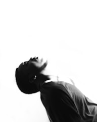 A black and white photograph of a person leaning back with their head tilted upward, displaying dramatic contrast between light and shadow over their face and shoulders. The image has an abstract and expressive feel, highlighting the contours and expressions subtly.