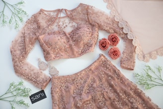 A detailed view of a delicate, peach-colored embroidered outfit, featuring a long-sleeved blouse and matching skirt adorned with intricate floral patterns. Beside the outfit, elegant earrings and artificial roses complement the ensemble. The setup is enhanced with lace fabric and green sprigs arranged artfully on a light surface.