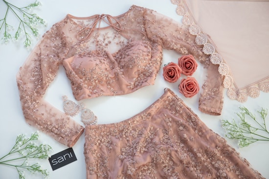 A detailed view of a delicate, peach-colored embroidered outfit, featuring a long-sleeved blouse and matching skirt adorned with intricate floral patterns. Beside the outfit, elegant earrings and artificial roses complement the ensemble. The setup is enhanced with lace fabric and green sprigs arranged artfully on a light surface.