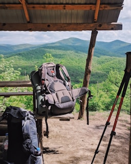 A wooden shelter overlooking a scenic view of lush, green mountains. Two hiking backpacks and trekking poles are prominently placed, suggesting a stop during a hiking trip. The open-sided shelter provides a shaded resting spot.