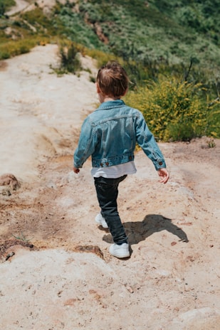 boy in green hoodie and blue denim jeans walking on brown dirt road during daytime