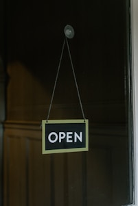 A simple open sign is hanging from a glass door with a suction cup. The sign has a black background with white text, bordered by a light color. The background consists of a dimly lit wooden wall panel.