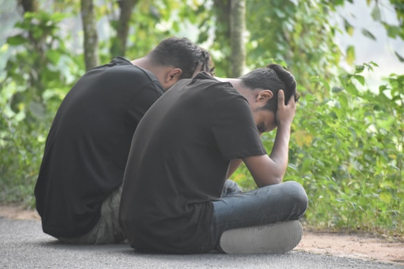 Two individuals are sitting on the side of a road surrounded by greenery. They are both wearing dark clothing, and their posture suggests they are experiencing sadness or contemplation. One person has their head in their hands.