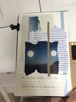 A collage on a notebook page features torn paper pieces arranged creatively. The background consists of printed text in various languages, layered with images of landscapes and celestial bodies. In the center, two white circles resembling moons are placed against a blue sky over mountains. Below, typed text on cream paper reads: 'May the space between where I am & where I want to be inspire me.' The composition is bordered by a black clip on the left edge.