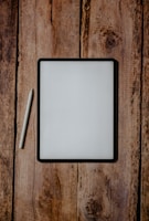 A digital tablet with a black frame rests on a rustic wooden surface, accompanied by a white stylus placed next to it on the left.