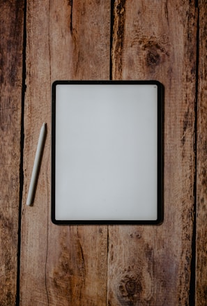 A digital tablet with a black frame rests on a rustic wooden surface, accompanied by a white stylus placed next to it on the left.