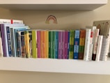 A collection of children's books neatly arranged on a white shelf. The books are organized by their size and color, ranging from multi-colored board books to classic children's stories. Above the books is a small rainbow sticker on the wall.