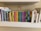 A collection of children's books neatly arranged on a white shelf. The books are organized by their size and color, ranging from multi-colored board books to classic children's stories. Above the books is a small rainbow sticker on the wall.