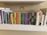A collection of children's books neatly arranged on a white shelf. The books are organized by their size and color, ranging from multi-colored board books to classic children's stories. Above the books is a small rainbow sticker on the wall.