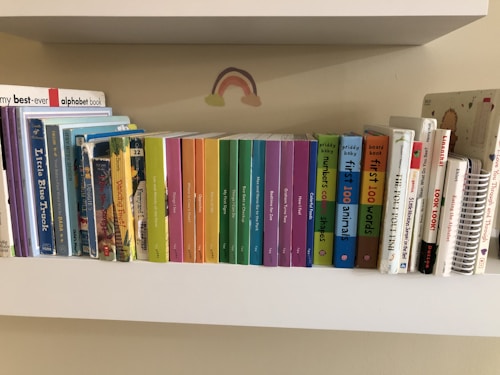 A collection of children's books neatly arranged on a white shelf. The books are organized by their size and color, ranging from multi-colored board books to classic children's stories. Above the books is a small rainbow sticker on the wall.
