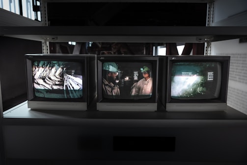 Three CRT monitors are displayed on a shelf, each showing different images. The left screen displays an industrial setting with machinery, the middle screen shows a person wearing a helmet, possibly a worker, and the right screen shows a blurred, abstract scene.