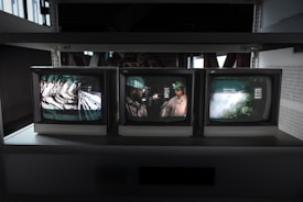 Three CRT monitors are displayed on a shelf, each showing different images. The left screen displays an industrial setting with machinery, the middle screen shows a person wearing a helmet, possibly a worker, and the right screen shows a blurred, abstract scene.