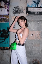 A young person stands against a concrete wall decorated with various posters, including a graphic design and a music-related advertisement. They are wearing a white tube top, white pants, and a belt with eyelets. A bright green handbag is slung over their shoulder.