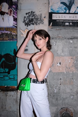 A young person stands against a concrete wall decorated with various posters, including a graphic design and a music-related advertisement. They are wearing a white tube top, white pants, and a belt with eyelets. A bright green handbag is slung over their shoulder.