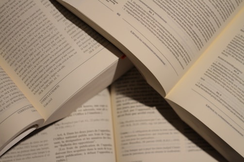 Several open books with visible text in a well-lit setting, showing pages filled with printed paragraphs. The books are arranged in a slightly overlapping manner.