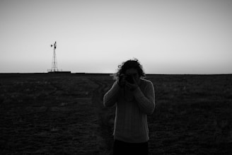 A black-and-white photograph captures a person in a sweater holding a camera against an open flat landscape. A windmill stands in the distance under a clear sky, creating a sense of isolation and calm.