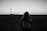 A black-and-white photograph captures a person in a sweater holding a camera against an open flat landscape. A windmill stands in the distance under a clear sky, creating a sense of isolation and calm.