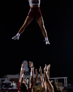 Cheerleaders perform a stunt at night. One cheerleader is airborne, captured mid-flight, supported by a group of teammates with their arms extended upwards. They are dressed in matching uniforms. The background is dark with the outline of a building visible.
