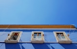 A vibrant blue building facade with three windows, each framed with a light yellow trim and decorative elements. The bottom half of the windows is guarded by black metal railings. Terracotta pots are visible around the windows. The vivid blue of the building contrasts sharply with the clear, bright blue sky above.