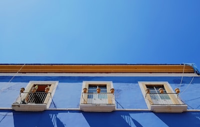 A vibrant blue building facade with three windows, each framed with a light yellow trim and decorative elements. The bottom half of the windows is guarded by black metal railings. Terracotta pots are visible around the windows. The vivid blue of the building contrasts sharply with the clear, bright blue sky above.
