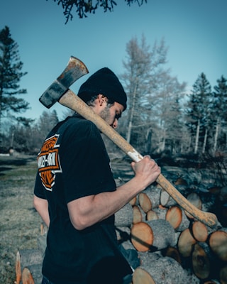 A person is standing outdoors in a forested area, holding an axe over their shoulder. They are wearing a black beanie and a black shirt with a visible logo on the back. In the background, cut logs are piled up, indicating wood chopping activity. The trees around are leafless, suggesting a cold season.