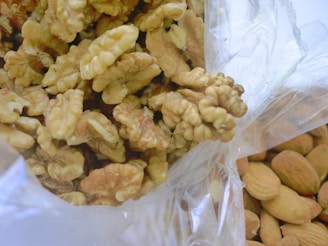 A pile of shelled walnuts is seen inside a transparent plastic bag. The nuts have a wrinkled texture and a light brown color. To the right, unshelled almonds are partially visible, showcasing their smooth, tan shells.
