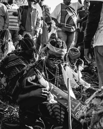 A group of people wearing traditional clothing and accessories, such as patterned blankets and beaded necklaces, are gathered outdoors. One individual in the foreground is wearing sunglasses and holds a stick, while the others are standing or sitting nearby.