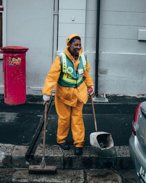 woman in green and yellow jacket holding black and brown stick standing beside red trash bin