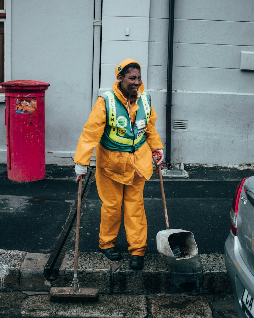 woman in green and yellow jacket holding black and brown stick standing beside red trash bin