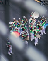 A vibrant marching band dressed in colorful, patterned costumes with white hats. They are playing various brass instruments like trombones, saxophones, and trumpets. One member holds a multicolored umbrella, adding to the lively scene on a city street.